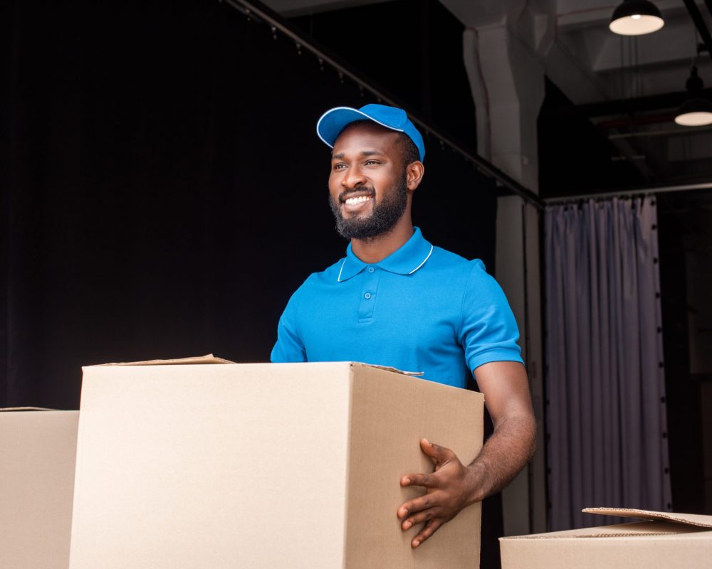 smiling african american delivery man holding box near storage